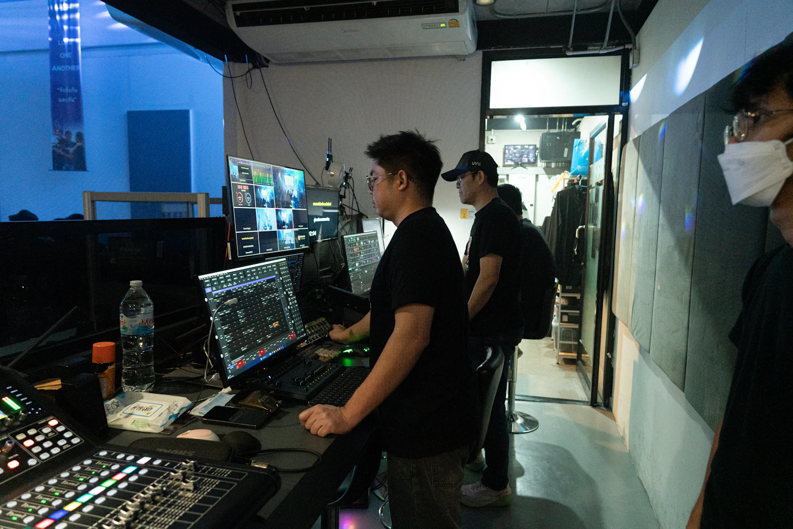 Production crew standing at a live video control desk with a multiview monitor, switcher panels and an audio mixing console in a dim room